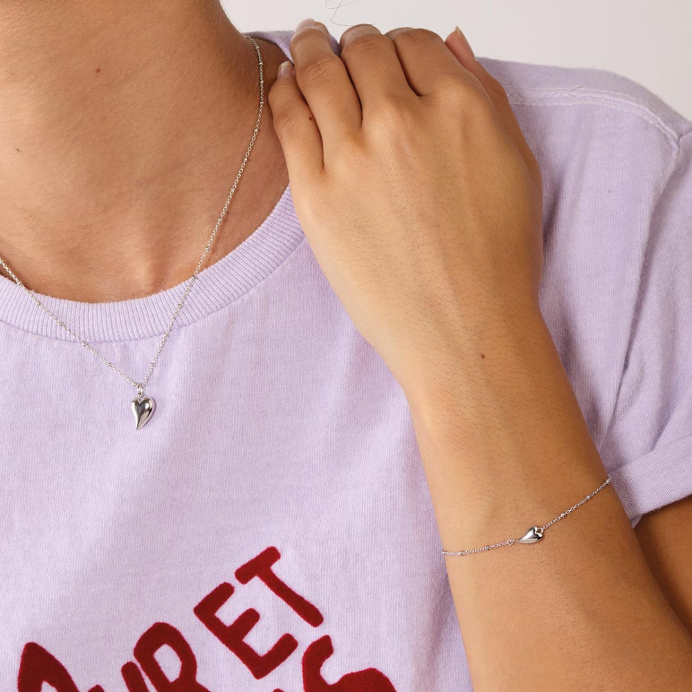 Person wearing a silver necklace and bracelet with heart pendants on a light purple shirt.