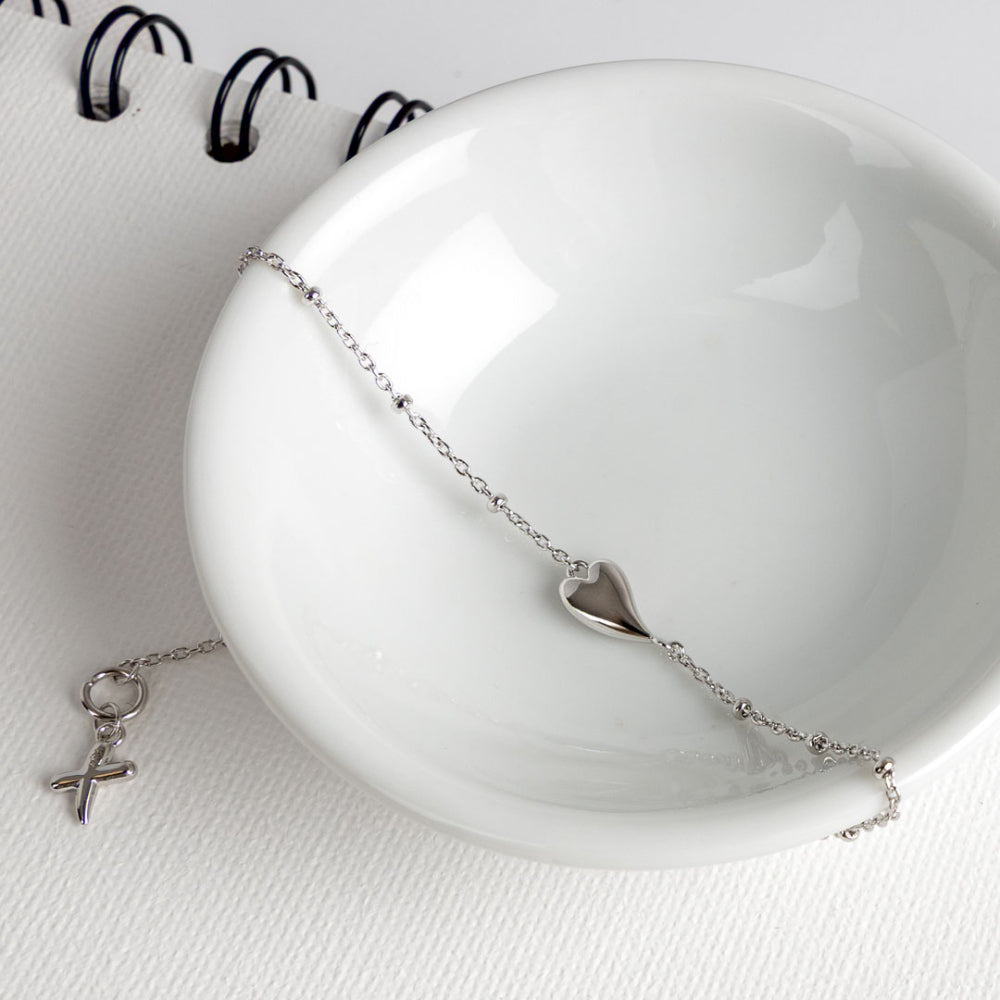 Silver bracelet with heart and cross charms on a white ceramic dish