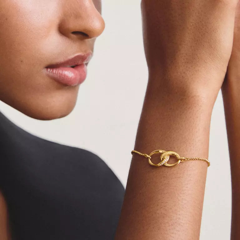 A close-up of a model's wrist wearing a delicate gold chain bracelet with two interlocking oval links, set against a neutral light gray background.
