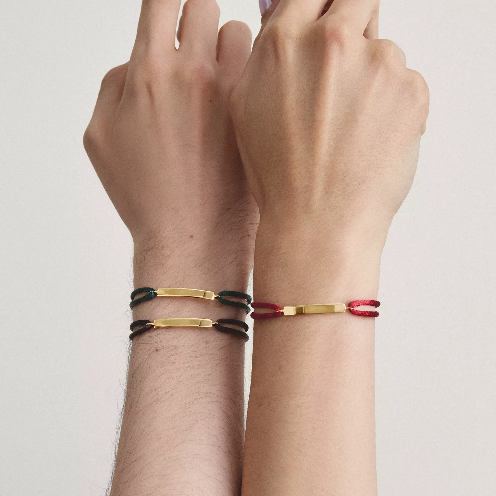 Two hands and wrists wearing slim metal bracelets with colored links. The left wrist has two bracelets, one black and one gold.