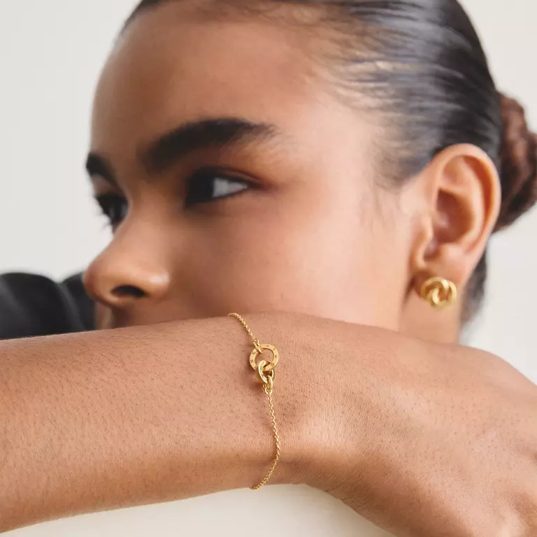 A delicate gold chain bracelet featuring a central interlocking double-ring charm, one of which is set with small clear stones. The bracelet includes an adjustable lobster clasp and a heart-shaped extender tag, all shown on a clean white background.