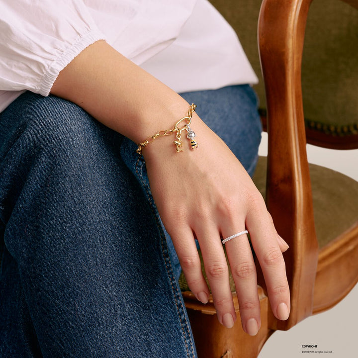 Hand wearing a gold bracelet with charms and a silver ring, sitting on a wooden chair.