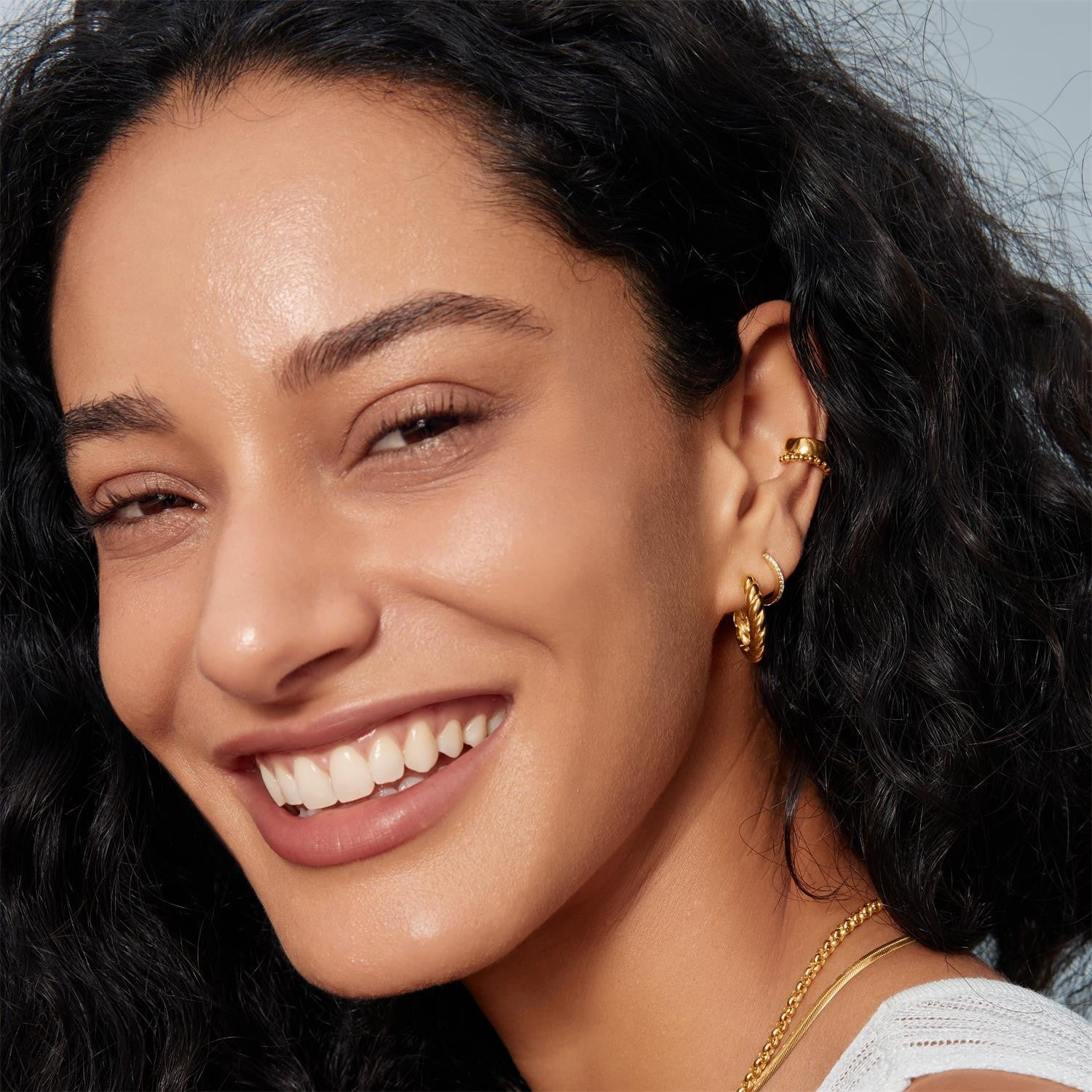 Close-up of a woman with dark hair and gold earrings, wearing a white top.