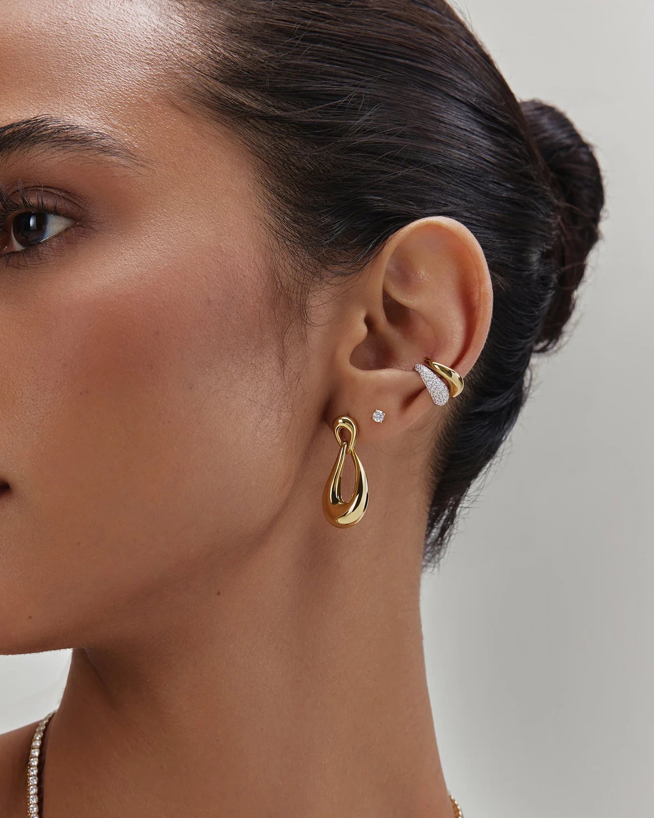 A close-up of a model's ear wearing a stack of gold and white stud earrings, including a large gold teardrop hoop, a small gold curved band, and a tiny white round stud.