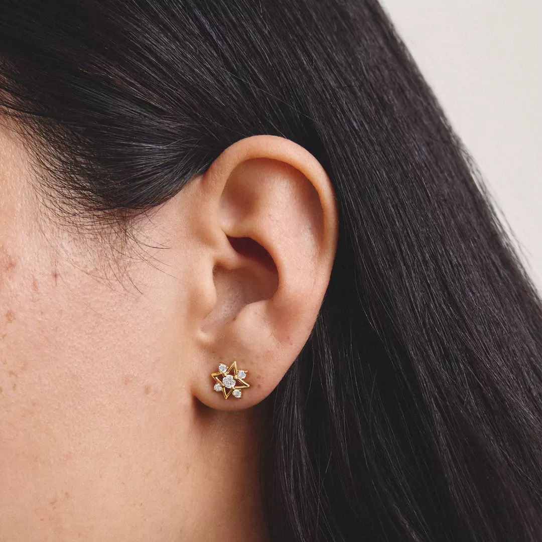 A close-up of a person's ear wearing a small gold stud earring with clear gemstones, against a light neutral background.