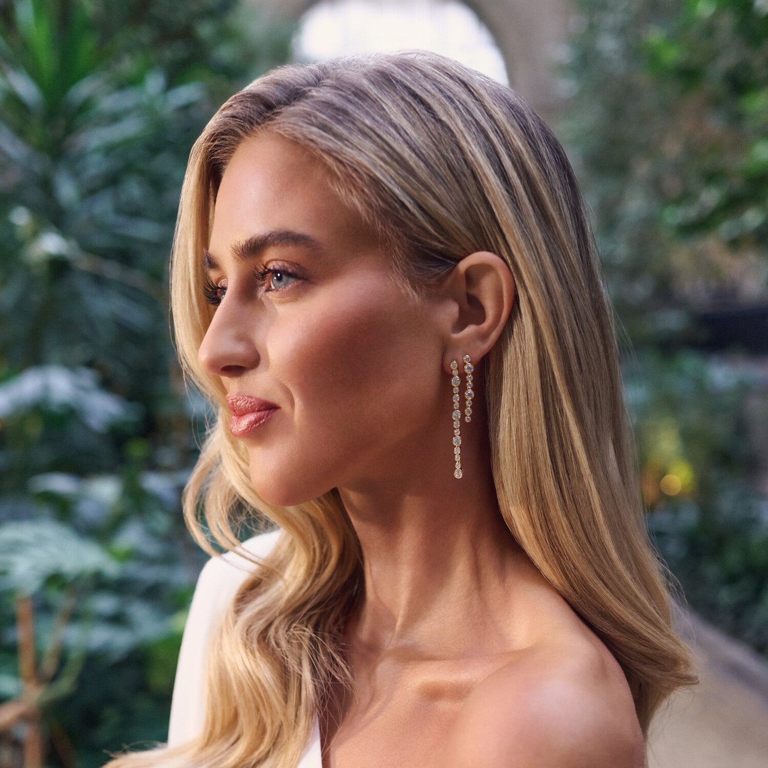 Woman with long blonde hair wearing earrings, with a blurred green outdoor background