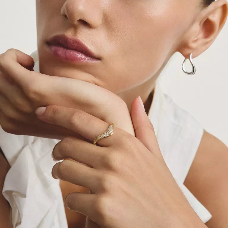 A close-up lifestyle shot of a model wearing a small, wavy diamond-encrusted gold ring on her finger, paired with a silver teardrop hoop earring and a white top.
