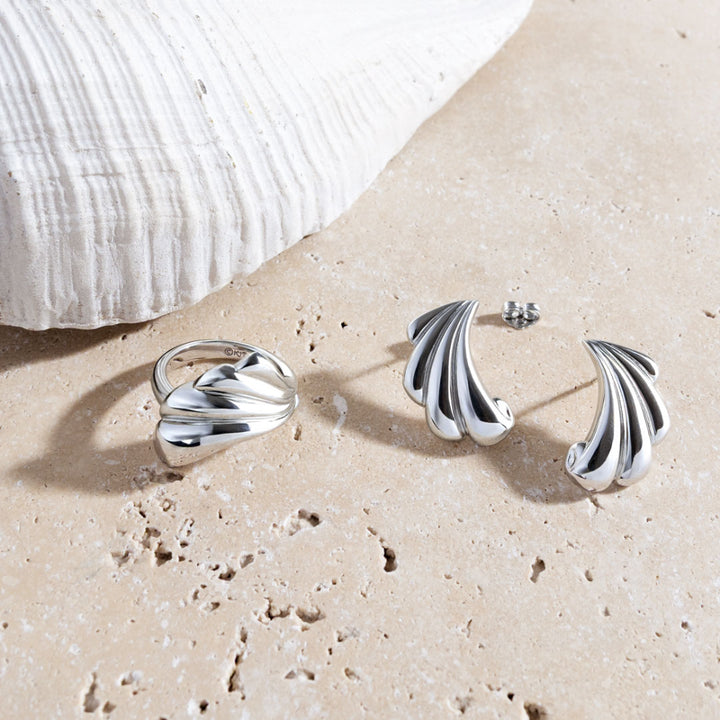 Silver jewelry set including a ring, earrings, and a necklace on sand with a shell in the background.