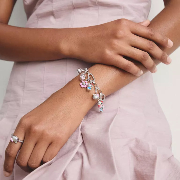 A silver charm bracelet with pink and light blue floral charms and pearl-like beads, shown worn on a woman's wrist. The woman is wearing a light pink dress, and a matching pink ring is also visible on her other wrist.