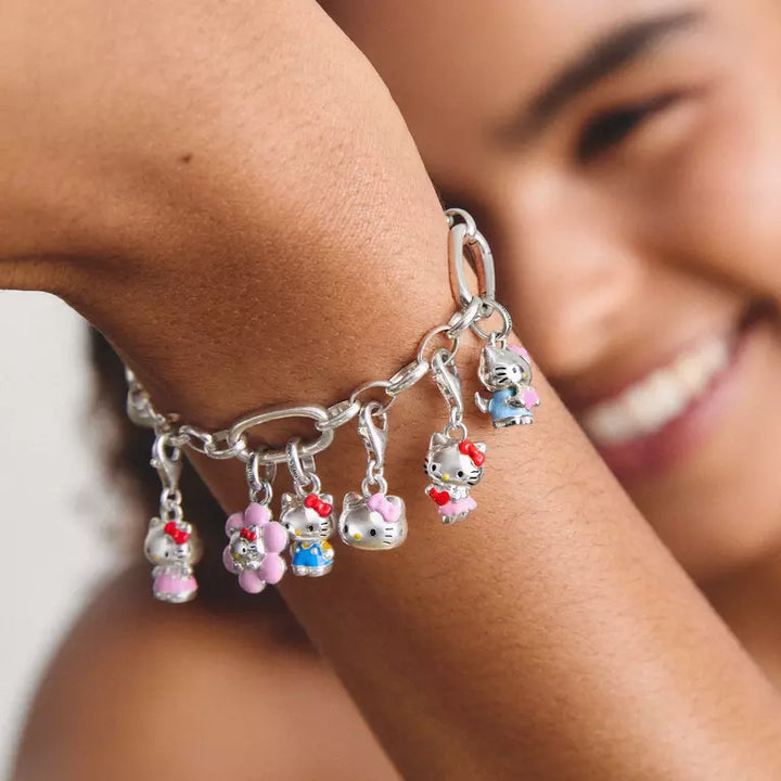 A close-up of a silver chain bracelet with multiple dangling Hello Kitty charms, worn on a person's wrist. The charms feature Hello Kitty's face and small flowers in pink, blue, and red, with a blurred smiling woman in the background.