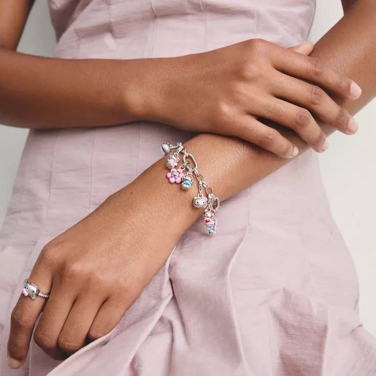 A silver charm bracelet with pink and light blue floral charms and pearl-like beads, shown worn on a woman's wrist. The woman is wearing a light pink dress, and a matching pink ring is also visible on her other wrist.