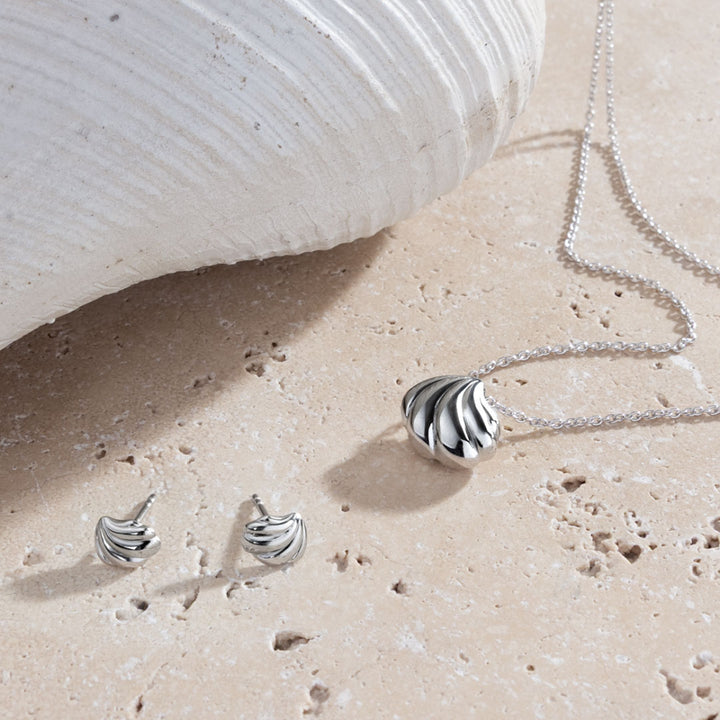 Silver jewelry set including necklace and earrings on a sandy surface with a shell in the background.