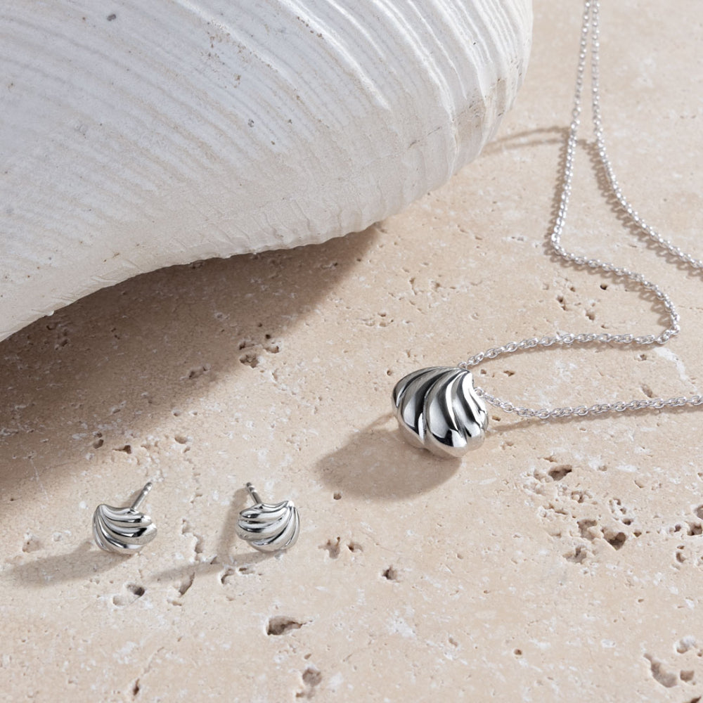 Silver jewelry set including necklace and earrings on a sandy surface with a shell in the background.