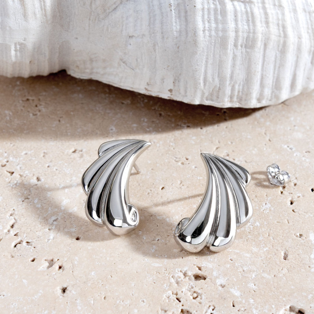 Silver shell-shaped earrings on a sandy surface with a shell in the background
