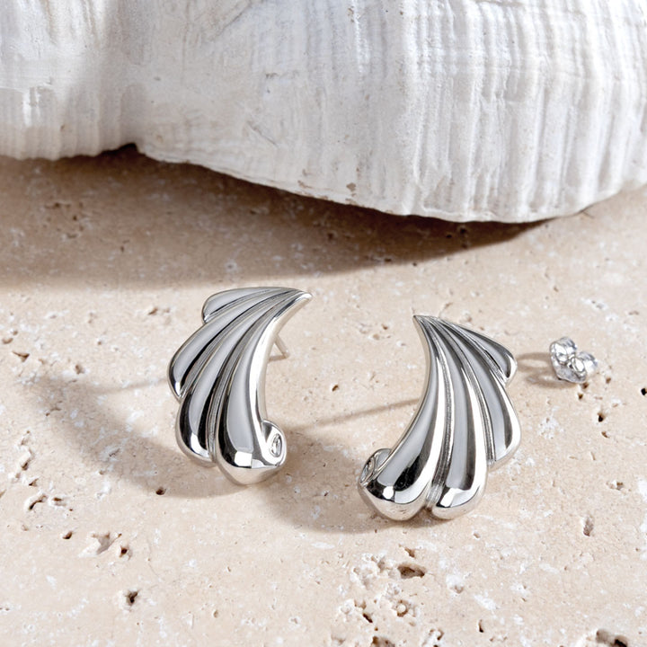 Silver shell-shaped earrings on a sandy surface with a shell in the background