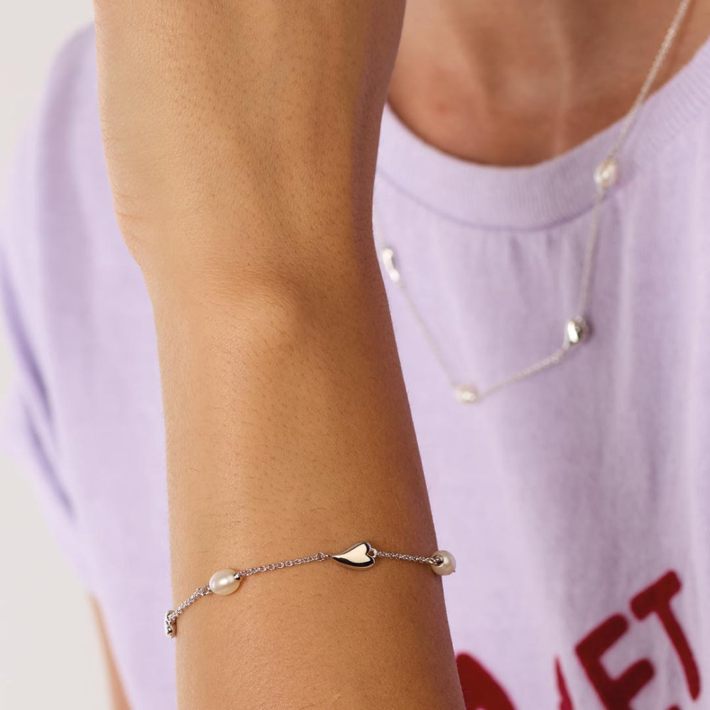 Close-up of a person's wrist wearing a delicate bracelet with a heart charm and pearl beads, against a blurred background.