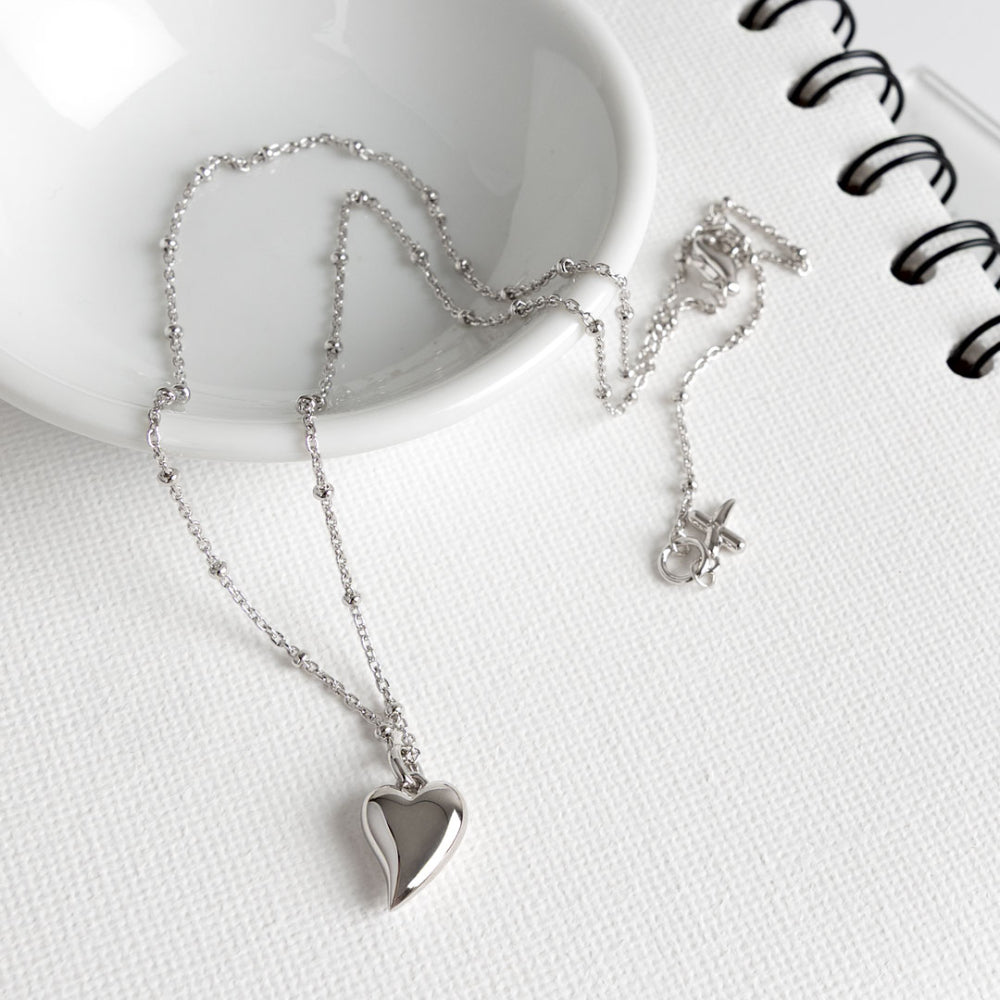 Silver heart-shaped pendant necklace on a white surface with a notebook.