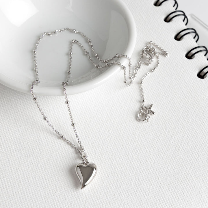 Silver heart-shaped pendant necklace on a white surface with a notebook.