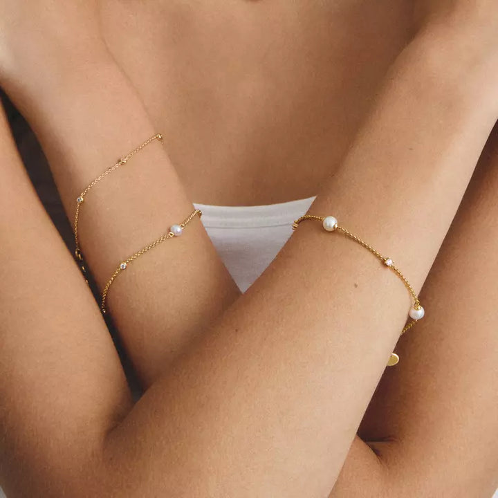 Two delicate gold chain bracelets with small white pearl accents worn on crossed forearms, shown on a model.