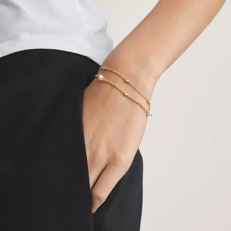 A close-up of a person's wrist wearing two delicate gold chain bracelets, one with a small pearl and the other with tiny clear stones, paired with a white top and black pants.
