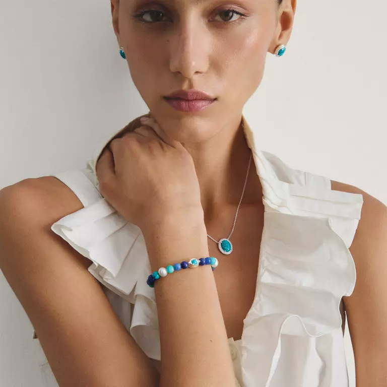 A close-up lifestyle shot of a model wearing a beaded bracelet and matching earrings with a blue pendant necklace, styled with a white ruffled blouse against a plain light background.