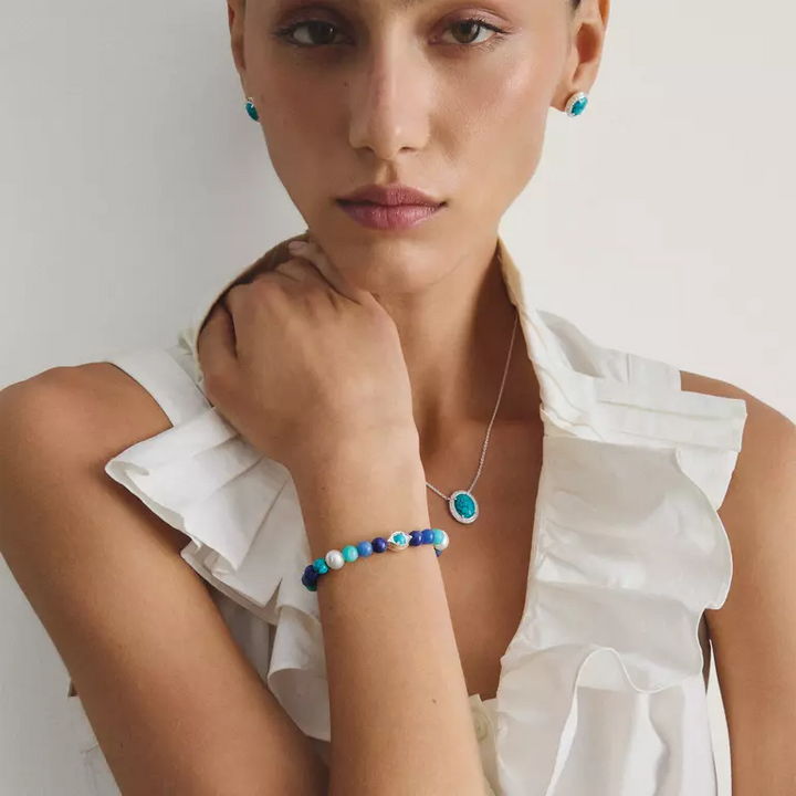 A close-up lifestyle shot of a model wearing a beaded bracelet and matching earrings with a blue pendant necklace, styled with a white ruffled blouse against a plain light background.