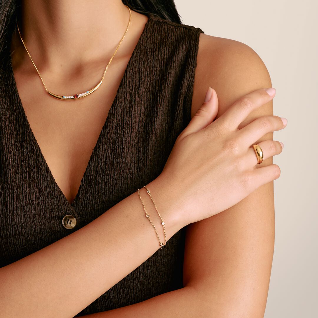 Woman wearing gold jewelry including a necklace, bracelet, and ring on a neutral background