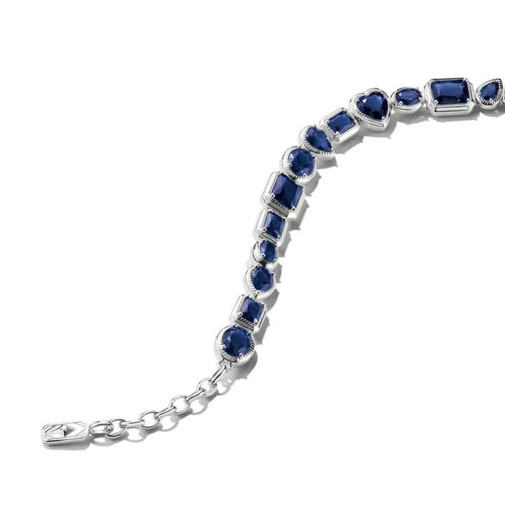 A silver-toned tennis bracelet featuring a series of deep blue gemstones in varied shapes, including emerald, round, and pear cuts, set in a silver-toned metal with a delicate chain extender clasp.