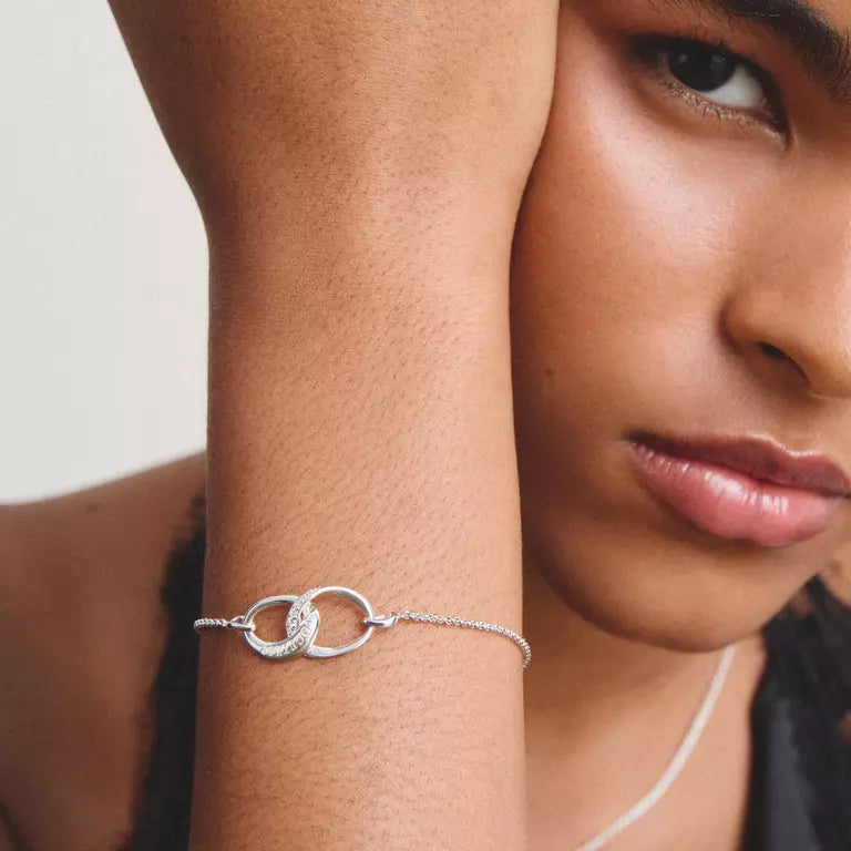 A close-up lifestyle shot of a model wearing a delicate silver chain bracelet with two interlocking oval rings on her wrist.