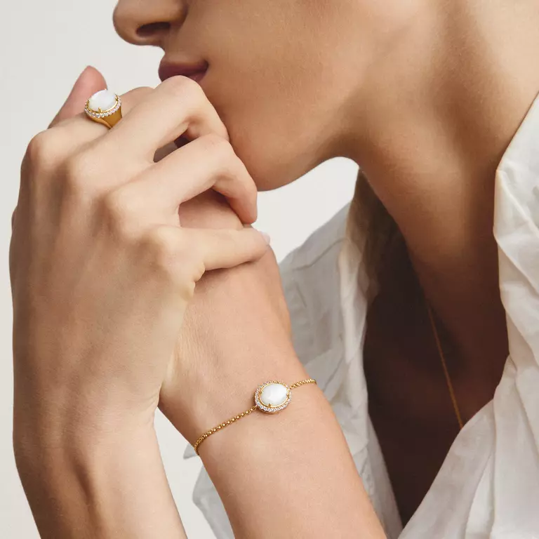 A close-up lifestyle shot of a model wearing a delicate gold chain bracelet with a round white pearl-like centerpiece, paired with a matching gold ring on her finger.