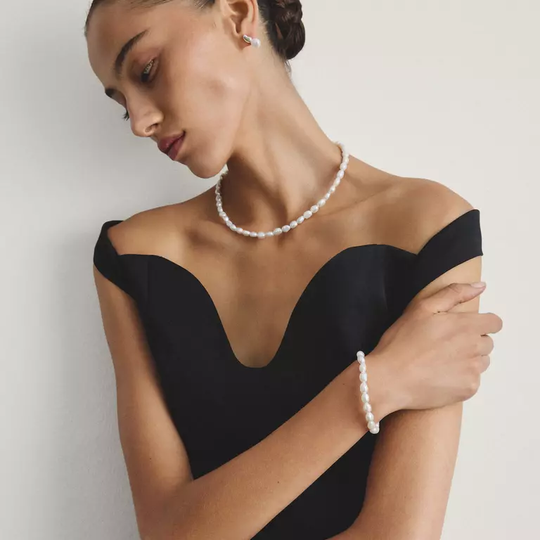 A woman wearing a pearl necklace and matching pearl bracelet, styled with a black off-the-shoulder dress against a neutral light gray background.