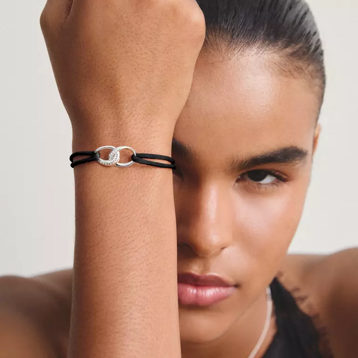 A close-up lifestyle shot of a model wearing a black double-wrap bracelet with a silver-toned interlocking rings design on her wrist.