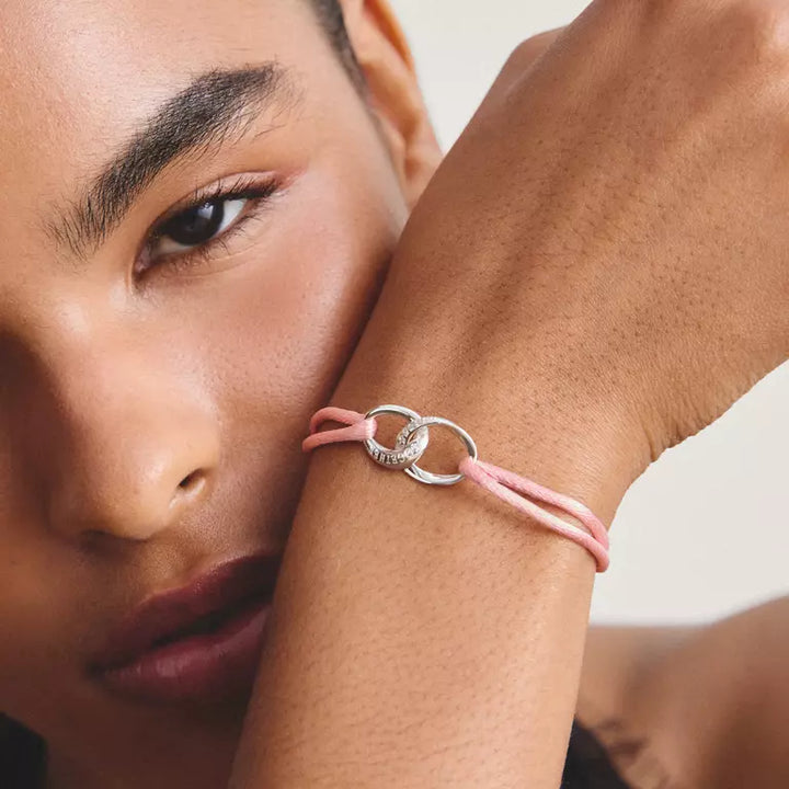 A close-up lifestyle shot of a model's wrist wearing a delicate pink cord bracelet with a silver interlocking rings clasp.