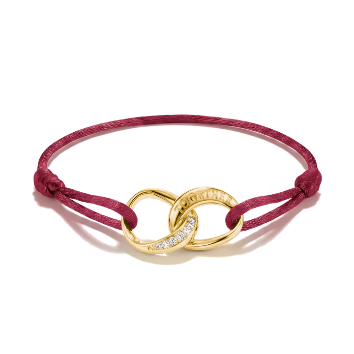 A burgundy cord bracelet with an interlocking gold infinity-style charm, accented with small clear stones and engraved with the brand name 'CHANEL'.