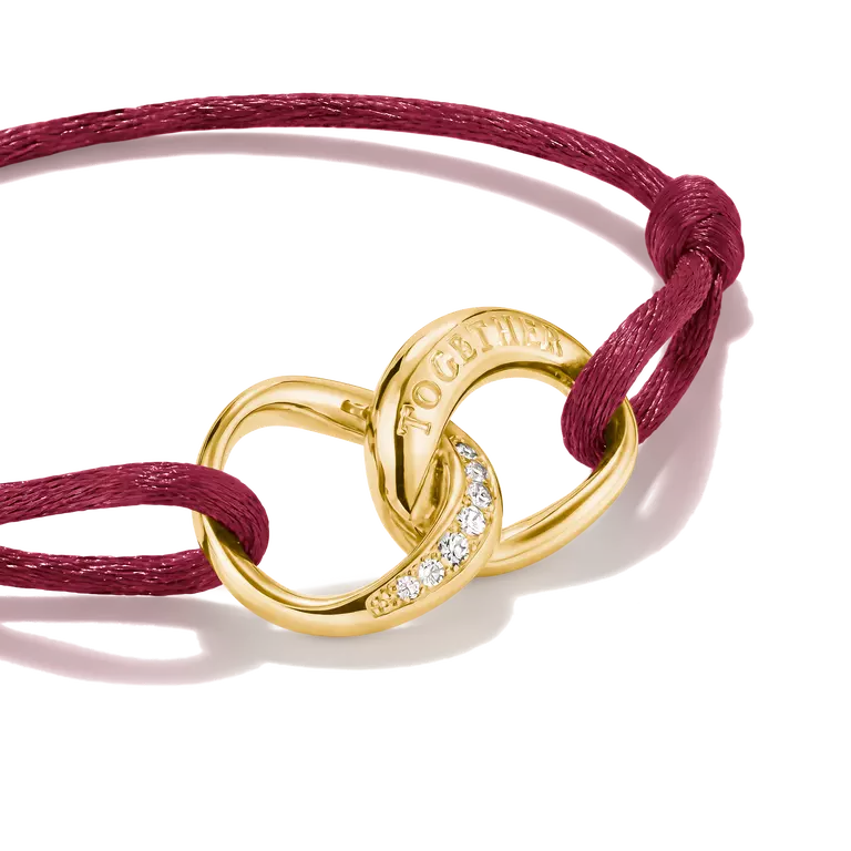 A close-up studio shot of a gold-toned interlocking knot bracelet with a row of small clear stones and the word 'TOGETHER' engraved on one loop, tied with a burgundy cord on a white background.