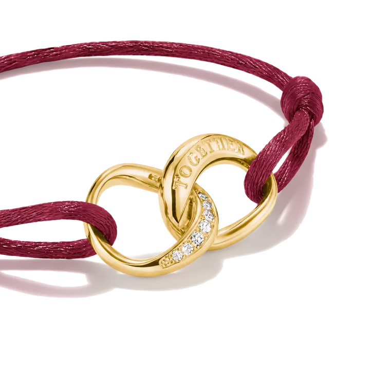 A close-up studio shot of a gold-toned interlocking knot bracelet with a row of small clear stones and the word 'TOGETHER' engraved on one loop, tied with a burgundy cord on a white background.