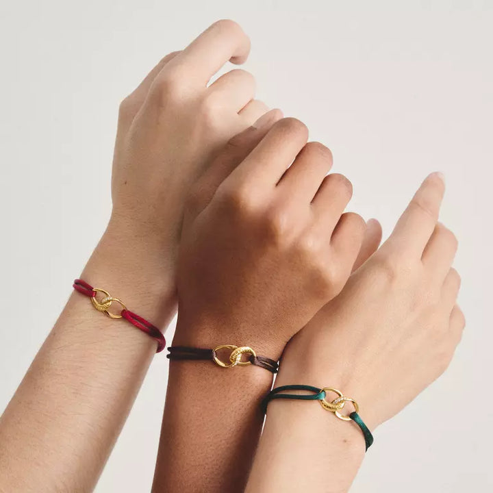 Three delicate cord bracelets with interlocking gold-tone rings are shown on three different arms against a light neutral background.