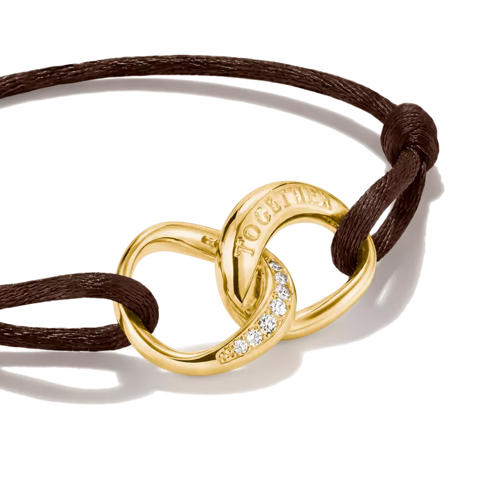 A gold knot bracelet with a dark brown cord and a row of small clear stones, shown on a white background.