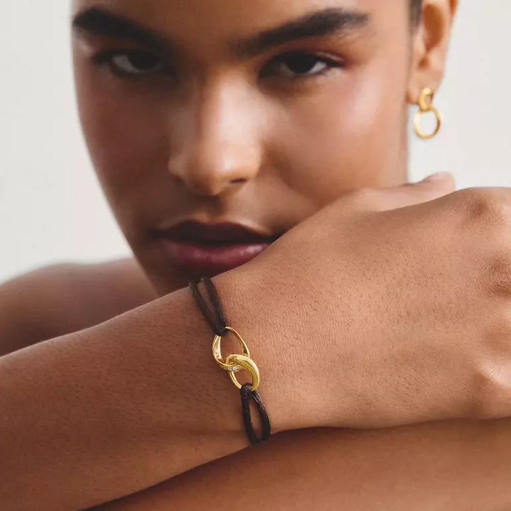 A close-up lifestyle shot of a model wearing a black cord bracelet with a gold interlocking rings design on her wrist, with a matching gold hoop earring visible on her ear.