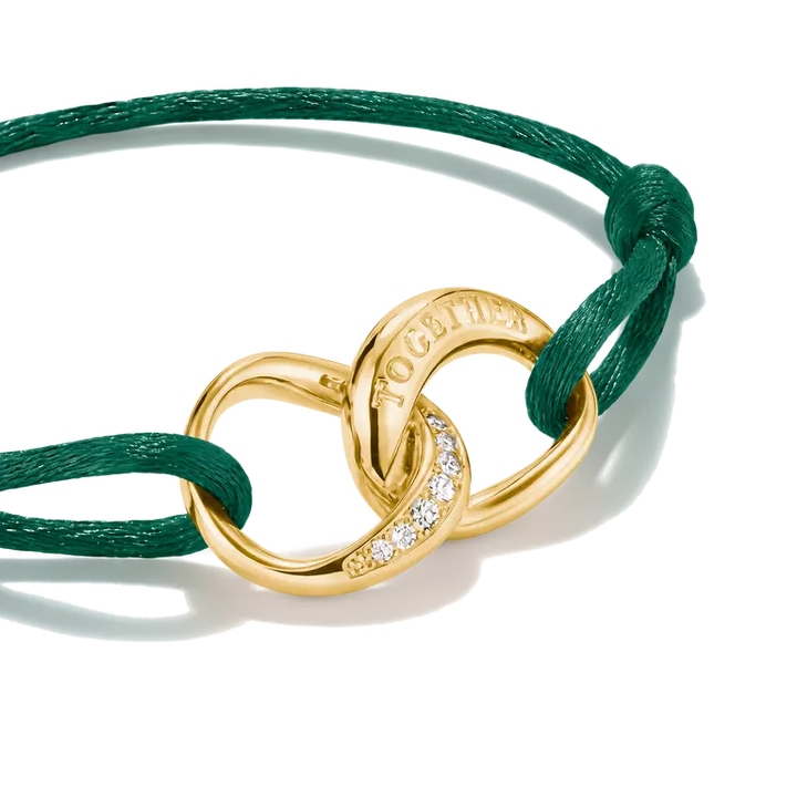 A gold-toned interlocking rings charm bracelet with a small row of clear stones and the engraved word 'TOGETHER', attached to a dark green cord on a white background.