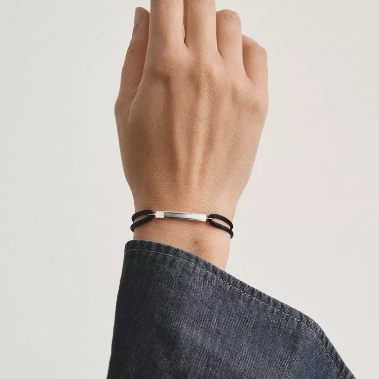 A close-up of a person's wrist wearing a minimalist bracelet with a polished silver bar and black cords, paired with a blue denim sleeve against a light gray background.