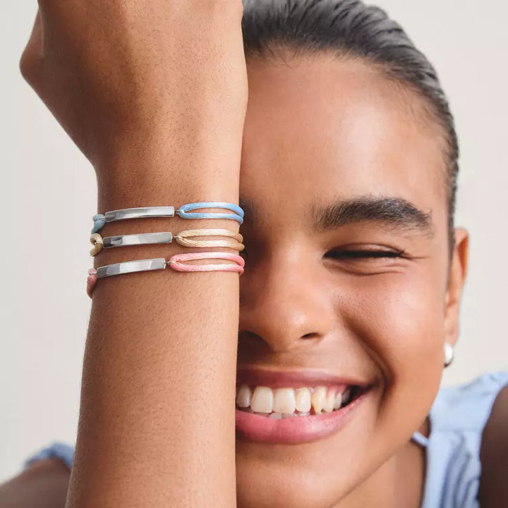 A close-up lifestyle shot of a smiling woman resting her head on her hand, which is adorned with four minimalist metallic bangles in silver, gold, and pink tones.