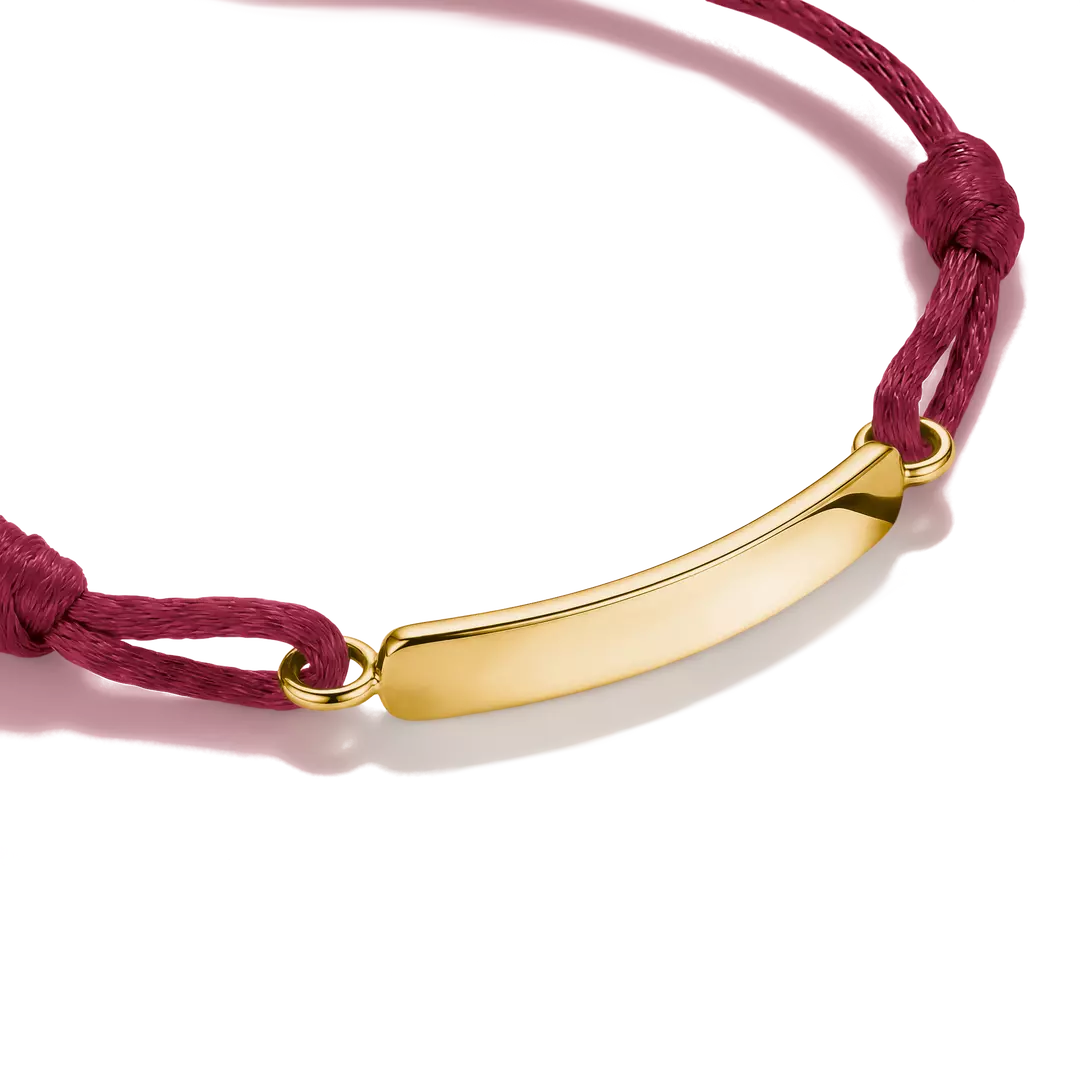 A close-up of a gold-plated curved bar nameplate bracelet on a deep red cord, set against a clean white background with a soft shadow.