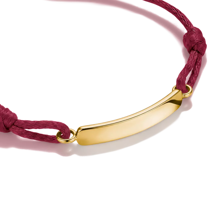 A close-up of a gold-plated curved bar nameplate bracelet on a deep red cord, set against a clean white background with a soft shadow.