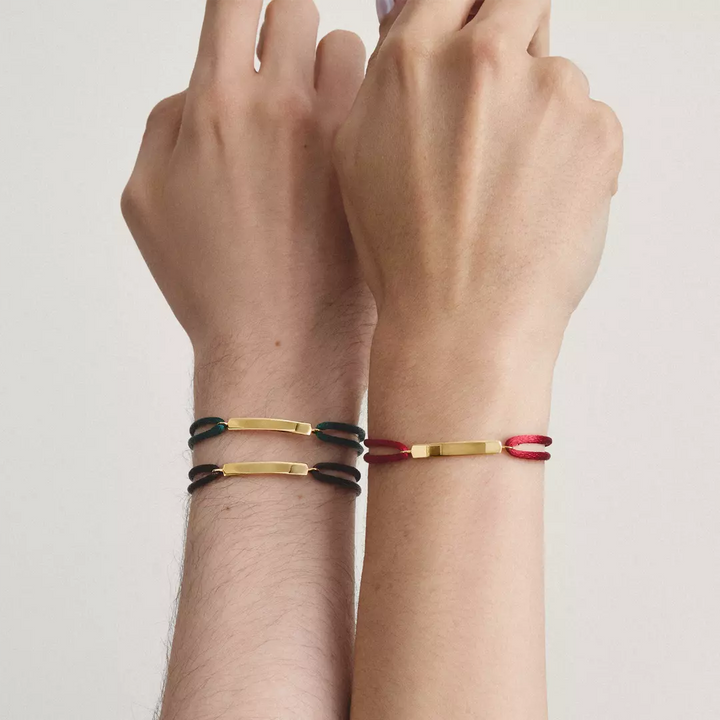 Two hands and wrists wearing slim metal bracelets with colored links. The left wrist has two bracelets, one black and one gold.
