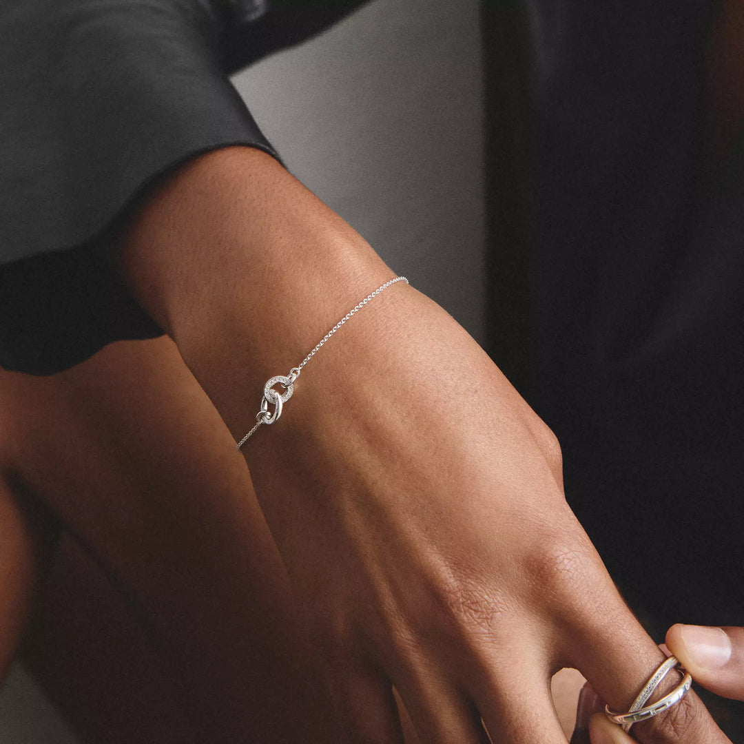 A delicate silver chain bracelet with a knotted charm, shown worn on a person's wrist. The person is wearing a black short-sleeve shirt, and a silver ring is visible on their ring finger.