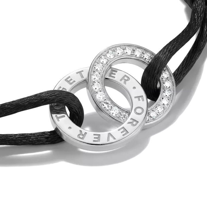 A close-up of a silver-toned interlocking rings pendant with clear pavé stones and engraved text reading 'FOREVER' and 'ET ER', threaded through a black cord on a white background.
