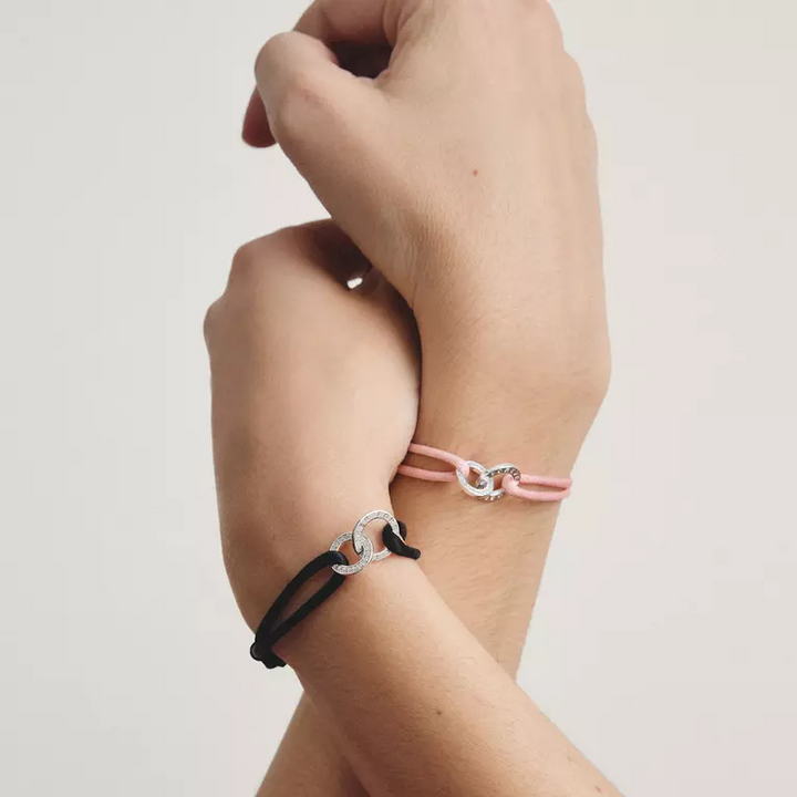 Two delicate bracelets shown on a person's wrist against a plain light background. One bracelet is black with a silver-toned interlocking ring motif, and the other is light pink with a similar silver-toned ring motif.