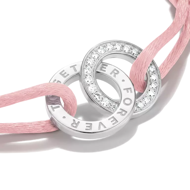 A close-up of a silver-toned double-ring charm bracelet with clear pavé stones and engraved text, threaded with a soft pink cord on a white background.