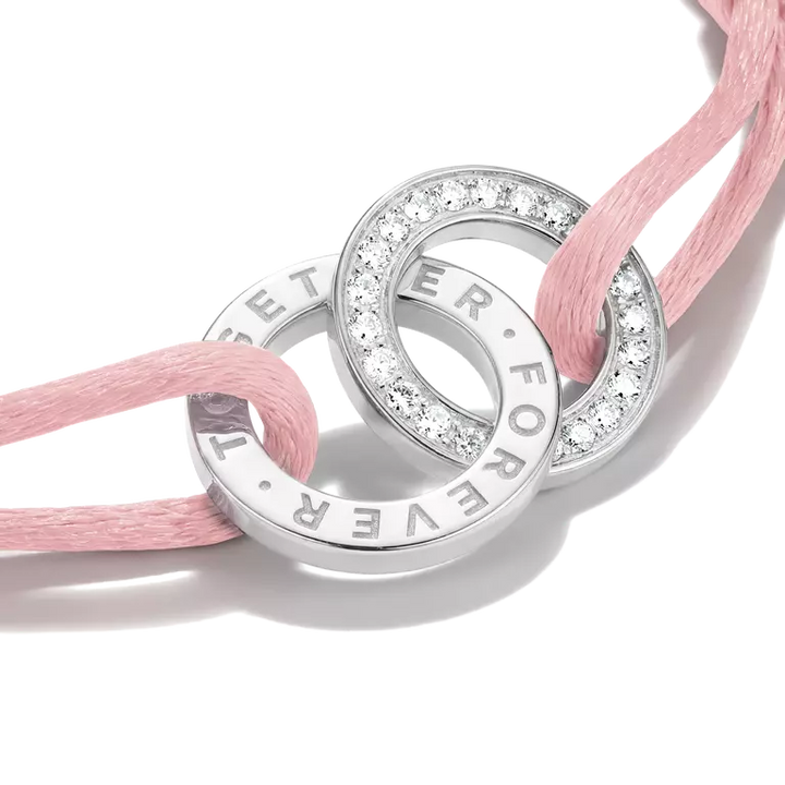 A close-up of a silver-toned double-ring charm bracelet with clear pavé stones and engraved text, threaded with a soft pink cord on a white background.